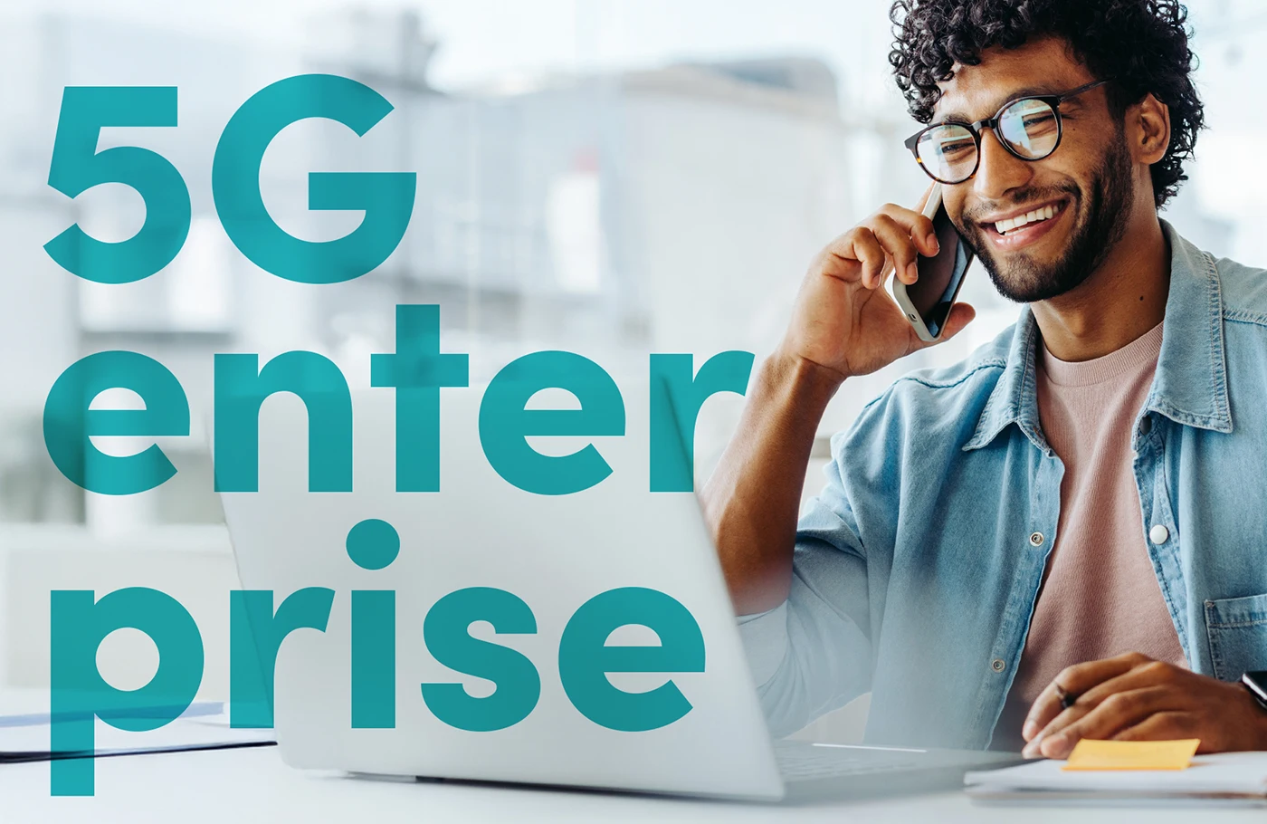 Lächelnder junger Mann mit Brille sitzt vor Laptop und hält sich ein Smartphone ans Ohr. Schriftzug 5G enterprise als Overlay.
